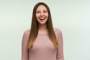 portret van jong vrouw met lang haar- omlaag, looks blij, lachend Aan een grap, lachend, gekleed in een strak roze breien trui, geïsoleerd over- wit achtergrond foto