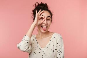 portret van grappig aantrekkelijk positief charmant brunette meisje met verzameld Golf haar- maken okey symbool dichtbij naar oog met een hand, shows tong, in blouse met polka stippen, geïsoleerd Aan roze achtergrond foto