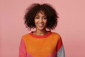 binnen- portret van positief optimistisch charmant Afrikaanse Amerikaans meisje met afro kapsel looks met genoegen, met levendig glimlach, vervelend kleurrijk lange mouw, geïsoleerd Aan roze achtergrond foto