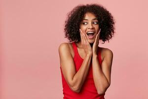 jong aantrekkelijk Afrikaanse Amerikaans met een afro kapsel met genot looks links naar de leeg ruimte, haar handen tintje haar gezicht, voelt verheugd, looks verrast, geïsoleerd Aan een roze achtergrond foto