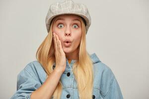 emotioneel blond jong vrouw met haar- omlaag, op zoek heel verrast met grote ogen, een hand- Holding haar wang, fluitjes in verrassing, draagt overmaats denim jasje, beige gecontroleerd pet, Aan grijs achtergrond foto