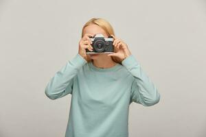 portret van een fotograaf aan het bedekken haar gezicht met de camera. fotograaf nemen foto, werken in studio, vervelend blauw gewoontjes trui, over- grijs achtergrond foto