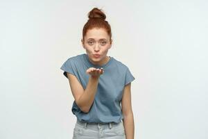 positief, flirterig vrouw met gember haar- broodje. vervelend blauw t-shirt. Bezig met verzenden lucht kus, blazen over- haar palm. emotie concept. aan het kijken lief Bij de camera, geïsoleerd over- wit achtergrond foto