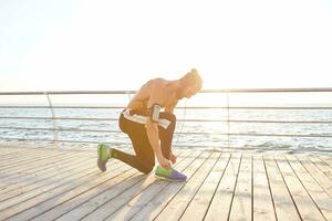 portret van jong knap sportief vent met baard, schaven rennen Bij de kust, luister favoriete mengen Aan koptelefoon. koppelverkoop schoenveters voordat joggen. foto