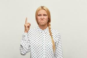 studio portret van een blond jong meisje met haar- verzameld in vlecht gefrustreerd verontrust van streek shows vinger omhoog lippen achtervolgd. iets mislukken naar vervullen haar hoopt of verwachtingen. over- wit achtergrond. foto