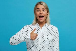 portret van gelukkig zelfverzekerd lief blond jong vrouw draagt polka punt overhemd glimlacht en shows duimen omhoog geïsoleerd over- blauw achtergrond foto