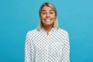 detailopname van vrolijk opgewonden schattig blond jong vrouw draagt polka punt overhemd voelt gelukkig en glimlachen tonen gezond tanden geïsoleerd over- blauw achtergrond foto