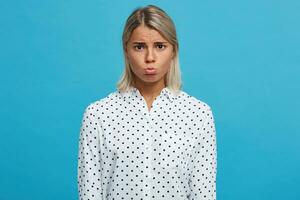 portret van verdrietig van streek mooi blond jong vrouw draagt polka punt overhemd voelt depressief en gebogen lippen geïsoleerd over- blauw achtergrond foto