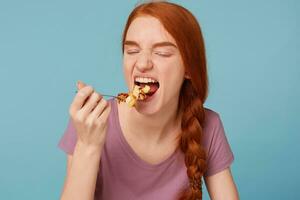 dichtbij omhoog van roodharig meisje Gesloten ogen, strekt zich uit lepel naar een mond eet een eet calorierijk taart, geïsoleerd Aan een blauw achtergrond foto