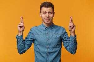 een jong Mens in een denim overhemd staat met zijn armen omhoog en vingers gekruiste looks Leuk vinden zwak geloven een wonder, maar nog steeds hopen voor mooi zo geluk. foto