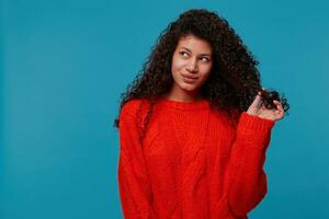 mooi spaans latino dame met gekruld donker haar- in rood gebreid trui Toneelstukken met een strand van haar- in haar hand, glimlachen dromend op zoek links kant Aan blanco kopiëren ruimte, geïsoleerd Aan blauw achtergrond foto