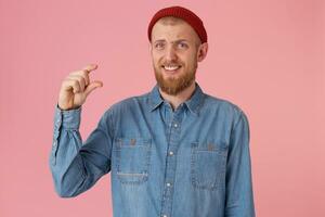 sceptisch gebaard mannetje in grappig rood hoed draagt denim shirt, maakt een hand- gebaar, demonstreert iets heel klein, poses tegen roze muur. verbaasd mannetje leerling shows hoe veel hij geleerd foto