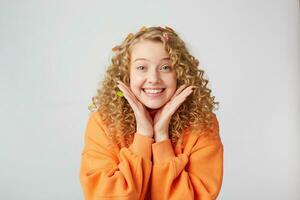 portret van lief inschrijving mooi hoor blond looks opgewonden, verrast, houdt palmen in de buurt gezicht, gekleed in te groot oranje trui, geïsoleerd Aan een wit achtergrond foto