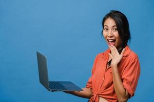 verraste jonge Aziatische dame met behulp van laptop met positieve uitdrukking, breed glimlachen, gekleed in casual kleding en camera kijken op blauwe achtergrond. gelukkige schattige blije vrouw verheugt zich over succes. foto