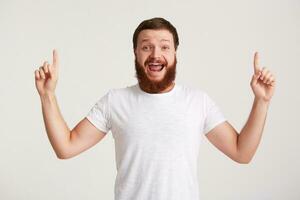 portret van bedrieglijk knap jong Mens hipster met baard draagt t overhemd voelt opgewonden en points omhoog Bij copyspace met vingers van beide handen geïsoleerd over- wit achtergrond foto