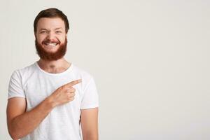 detailopname van glimlachen aantrekkelijk jong Mens hipster met baard draagt t overhemd voelt gelukkig en points naar de kant Bij copyspace met vinger geïsoleerd over- wit achtergrond foto