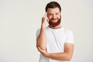 portret van glimlachen nadenkend knap jong Mens met baard draagt t overhemd houdt handen gevouwen, aanraken zijn tempel en denken geïsoleerd over- wit achtergrond foto