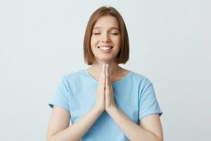 gelukkig vredig mooi jong vrouw in blauw t overhemd houden handen gevouwen in bidden positie en ogen Gesloten geïsoleerd over- wit achtergrond glimlachen en mediteren foto