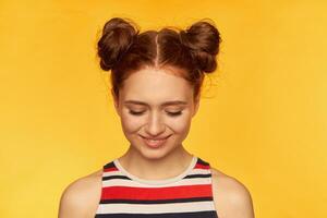 tiener- meisje, gelukkig op zoek, charmant rood haar- vrouw met twee broodjes. vervelend gestreept overhemd en aan het kijken naar beneden met glimlach, verlegen. detailopname, staan geïsoleerd over- geel achtergrond foto