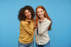 blij aantrekkelijk Dames tonen hun wit perfect tanden terwijl glimlachen breed Bij camera, houden palmen verheven terwijl poseren over- blauw achtergrond en knuffelen elk andere foto