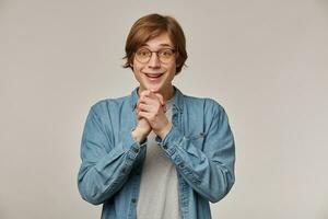 portret van vrolijk, positief mannetje met blond haar. vervelend denim shirt, bril en heeft een beugel. houdt zijn palmen samen en lachend. aan het kijken Bij de camera geïsoleerd over- grijs achtergrond foto