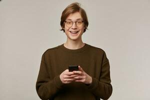 portret van knap, vrolijk mannetje met blond haar. vervelend bruin trui, bril en heeft een beugel. Holding een smartphone en lachend. aan het kijken Bij de camera geïsoleerd over- grijs achtergrond foto