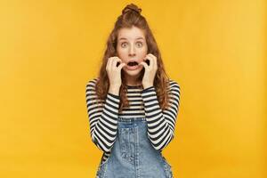 binnen- schot van jong vrouw met een lang rood haar, draagt denim overall en gestript shirt, met in de hoofdrol in camera met bang gelaats uitdrukking terwijl aan het kijken verschrikking film. foto