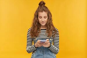 binnen- studio schot van jong gember vrouw met lang rood gekruld haar- houden kalmte terwijl typen bericht Aan haar telefoon geïsoleerd over- geel achtergrond foto