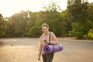 horizontaal foto van jong schattig dame wandelen na yoga Bij de park en chatten verstand vriend, hols de smartphone in hand- en looks Bij de Scherm met glimlach, luisteren muziek- in koptelefoon.