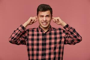 portret van jong aantrekkelijk vent in geruit shirt, looks Bij de camera, staat over- roze achtergrond en covers oren van luid lawaai. foto