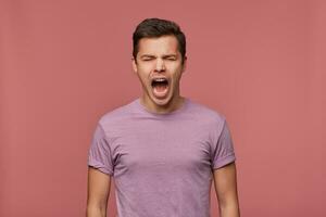 foto van jong aantrekkelijk vent in blanco t-shirt, staat over- roze achtergrond met Gesloten ogen en schreeuwen, looks boos en ongelukkig.