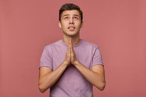 jong aantrekkelijk vent draagt in blanco t-shirt, maken een wens, hoopt voor mooi zo geluk en looks omhoog, shows gebeden gebaar, staat over- roze achtergrond. foto