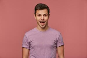 portret van vrolijk knap jong Mens draagt in blanco t-shirt, looks Bij de camera met gelukkig uitdrukking, staat over- roze achtergrond. foto