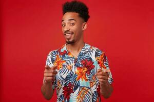 jong positief Afrikaanse Amerikaans Mens draagt in hawaiiaans shirt, looks Bij de camera en breed glimlacht, shows vingers naar de camera, staat over- rood achtergrond. foto