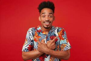 portret als jong positief Afrikaanse Amerikaans Mens in hawaiiaans shirt, looks Bij de camera met vrolijk uitdrukking, staat over- rood achtergrond en breed glimlacht, points in verschillend routebeschrijving. foto