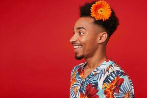 portret van jong gelukkig donker huid jongen, draagt in hawaiiaans shirt, looks weg met gelukkig uitdrukking, met een oranje bloem in haar, staat over- rood achtergrond. foto