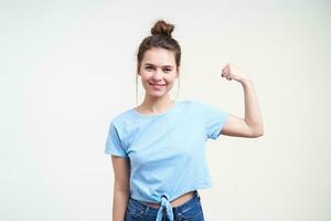 vrolijk jong lief brunette dame met gewoontjes kapsel glimlachen graag terwijl tonen haar sterk biceps, staand over- wit achtergrond in blauw t-shirt foto