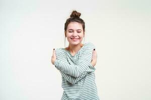 verheugd jong lief bruin haren dame houden haar ogen Gesloten terwijl knuffelen haarzelf en glimlachen aangenaam, staand over- wit achtergrond in gestreept blouse foto