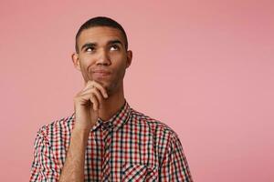 dichtbij omhoog van jong nadenkend aantrekkelijk donker huid vent in geruit shirt, looks weg en accenten kin, denkt en twijfels, staat over- roze achtergrond met kopiëren ruimte Bij de Rechtsaf kant. foto