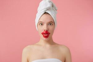 portret van jong grappig vrouw met lap voor lippen, na douche met een handdoek Aan haar hoofd, looks Bij de camera, shows tong, staat over- roze achtergrond. foto