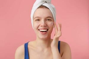 dichtbij omhoog van jong mooi hoor glimlachen vrouw met natuurlijk schoonheid met een handdoek Aan haar hoofd na douche, op zoek Bij de camera over- roze achtergrond en zet Aan gezicht room. foto