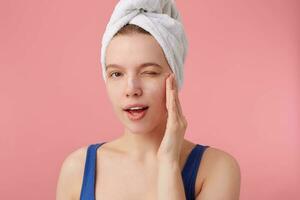 dichtbij omhoog van denken jong vrouw met een handdoek Aan haar hoofd na douche, kan niet maken een beslissing, looks Bij de camera en knipoog, accenten wang, staat over- roze achtergrond. foto