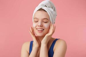 dichtbij omhoog van jong mooi hoor blij vrouw met natuurlijk schoonheid met een handdoek Aan haar hoofd na douche, staat over- roze achtergrond en zet Aan gezicht room met Gesloten ogen. foto