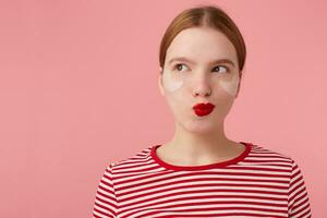 aantrekkelijk jong mysterieus roodharig vrouw met rood lippen en met patches onder de ogen, draagt in een rood gestreept t-shirt, looks Bij de links kant, iets plotten, staat over- roze achtergrond. foto