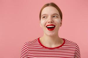 dichtbij omhoog van aantrekkelijk jong gelukkig roodharig vrouw met rood lippen en met patches onder de ogen, draagt in een rood gestreept t-shirt, looks weg en breed lachend, staat over- roze achtergrond. foto