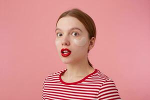 portret van schattig jong verbaasd roodharig dame in een rood gestreept t-shirt, met rood lippen en met patches onder de ogen, angstig looks Bij de camera, staat over- roze achtergrond. foto