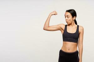 versteld staan jong slank brunette vrouw met paardenstaart kapsel op zoek verbaasd Aan haar biceps terwijl poseren over- wit achtergrond in zwart sportief beha top en leggins foto