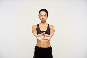 geconcentreerd jong bruine ogen sportief vrouw met gewoontjes kapsel kruispunt haar vingers terwijl uitrekken handen, aan het doen ochtend- training terwijl staand over- wit achtergrond foto