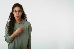 verbijsterd jong lang haren gekruld donker huid vrouw in eyewear fronsen verward wenkbrauwen terwijl tonen terzijde met inhoudsopgave vinger, poseren over- wit achtergrond foto