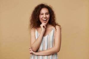 portret van jong lief gekruld brunette vrouw geven knipoog en tonen vrolijk tong terwijl op zoek Bij camera, houden hand- verheven terwijl poseren over- beige achtergrond foto